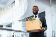 © Prostock-studio - A man wearing a suit smiles while holding a box filled with personal items. He stands in a modern office space during the day. The atmosphere appears bright and welcoming.