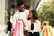 © Prostock-studio - Couple In Love. Cheerful handsome African American guy hugging his girlfriend or wife who holding colorful shopping bags and looking at him. Happy black lovers having fun outdoors and laughing