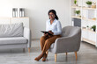 © Prostock-studio - Full length portrait of friendly black female psychologist with clipboard sitting in cozy armchair at modern office, copy space. Confident psychotherapist smiling at camera in mental health clinic