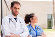 © StockMe - A serious-looking doctor and a nurse with stethoscopes and professional uniforms. Standing with their arms crossed, in front of a brightly lit health center.