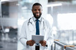 © Prostock-studio - A doctor stands in a modern healthcare facility with a tablet in hand. He is smiling and wearing a white coat with a stethoscope around his neck during work hours.