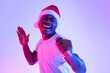 © Prostock-studio - A young black man wearing a Santa hat joyfully sings karaoke into a microphone. He dances and enjoys himself at a Christmas party, surrounded by colorful neon lights, creating a festive atmosphere.