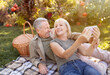 © Prostock-studio - Modern elderly spouses taking selfie on smartphone, having picnic and resting in garden at sunny autumn day, copy space. Senior couple capturing happy moments
