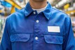 © Nanda - A worker wears a blue uniform shirt with a blank name tag patch. Use to represent occupations like manufacturing, repair, or warehouse jobs.