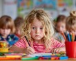 © patiporn - A young blonde girl with curly hair is drawing with a pencil at a table in a colorful kindergarten classroom surrounded by classmates.