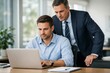© imagemir - A focused young Caucasian man works on a laptop while an older Caucasian man offers guidance in a modern office setting.