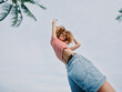 © SHOTPRIME STUDIO - Playful portrait of a smiling young woman jumping outdoors, wearing a striped crop top and denim shorts, captured from a low angle against a bright sky.
