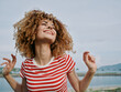 © SHOTPRIME STUDIO - A joyful woman with curly hair smiles toward the sky, wearing a red striped t-shirt, standing by a calm shore with distant hills and soft blue sky.