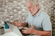 © luciano - Senior man sitting at desk in home office, looking stressed and frustrated while using laptop. Concept of technology problems, online difficulties, stress, remote work, aging and modern lifestyle