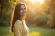 © miss irine - Smiling woman with long brown hair stands in a park during golden hour. Backlit by the sun she looks to the side with a happy expression. She wears a yellow sweater and enjoys nature.