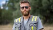 © Bussakon - A man with a beard and sunglasses wears a grey high visibility vest with yellow reflective stripes looking seriously at the outdoors