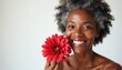 © Viktor - Smiling middle aged african american woman holds red flower near face. Graying curly hair, radiant skin, posing against white background. Happy expression conveys confidence, natural beauty in mature