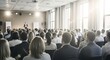 © Karmiyem - Professional business conference with a large audience attending a presentation in a modern conference room with natural sunlight streaming through large windows and speakers at the front