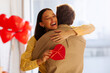 © Home-stock - Happy woman smiling and hugging man while holding heart-shaped gift box, red balloons fill the room, couple celebrating anniversary or Valentine's day