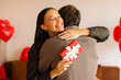 © Home-stock - Happy woman hugging man while holding gift behind his back, posing in room with red heart decorations, enjoying romantic moment, celebrating Valentine's Day