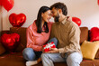 © Home-stock - Romantic couple celebrating Valentine's Day at home, bonding, man giving gift box to happy wife, sitting on sofa in decorated room