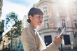 © Marko Geber - Adult woman smiling while using smartphone on sunny city street