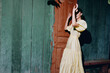 © SHOTPRIME STUDIO - A woman in a pale yellow dress stands beside a green wall and a wooden door, posing with her hands raised, sunlight casting a bold shadow in a vintage outdoor scene.