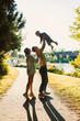 © DSMT - Family enjoying a sunny day by the riverside with a child being lifted in joy while walking on a path lined with greenery