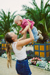 © DSMT - Mother lifts her child in a garden with palm trees and flowers during the day