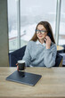 © DSMT - Woman talks on phone while sitting at table with laptop and coffee cup during winter season in modern indoor setting