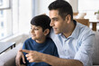 © fizkes - Indian father and little son sitting on couch at home, gazing out window with interest and curiosity, dad points at something outside, engaged in conversation indoors. Nurture, upbringing, fatherhood
