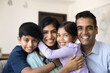 © fizkes - Close up portrait of loving Indian parents with cute little kids hugging seated on sofa at home, posing for photo, looking at camera, celebrate happy family day. Parenthood, upbringing, relationships