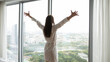 © fizkes - Triumph over the city. Rear view excited young female in robe stand by floor to ceiling window with wide open arms celebrate success of buying own high rise apartment spending vacation in luxury hotel