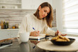 © Impact Photography - Woman writing notes in planner and using tablet computer while working remotely from home