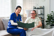 © PRIME STOCK LAB - Asian female nurse in blue scrubs showing a medical folder to a smiling senior man during a home healthcare visit in a living room.