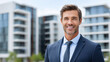 © Zanna - Confident businessman smiling outdoors in modern urban residential district, professional real estate agent standing in front of contemporary apartment buildings, success career concept in property