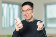 © fotolite - A smiling Chinese student wearing glasses holds a smartphone and credit card while making an online purchase, perfect for mobile payment, e-commerce, or student finance concepts.