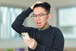 © fotolite - A frustrated Chinese student wearing glasses scratches his head in confusion while looking at his smartphone with a worried expression, ideal for financial stress, or other online problems.