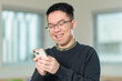 © fotolite - A happy Chinese student wearing glasses smiles while using his smartphone, perfect for mobile technology, social media, or student lifestyle concepts.