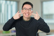 © fotolite - A cheerful Chinese student wearing glasses points to his bright smile with both index fingers, perfect for dental care, orthodontics, or oral health concepts.