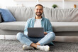 © Prostock-studio - Full length of happy young guy sitting cross legged on floor, using laptop computer, smiling at camera indoors. Positive millennial freelancer working online from home, attending business webinar