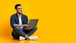 © Prostock-studio - Happy arab young man sitting on floor, using new modern laptop, typing on keyboard and smiling over yellow studio background, looking at copy space for text or advertisement, panorama