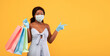 © Prostock-studio - Shopping during coronavirus. Young black lady in summer dress holding gift bags, wearing face mask and rubber gloves, pointing at empty space on orange background, panorama