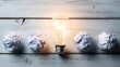 © Pixel Stone - Bright glowing incandescent light bulb shines triumphantly between several crumpled rejected paper balls representing failed ideas and new inspiration beginning