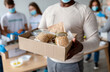 © Prostock-studio - A senior black man holds a donation box filled with various food items such as grains, canned goods, and pasta. In the background, millennial volunteers pack more boxes to help those in need.