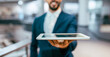 © Prostock-studio - A businessman holds a tablet out in front of him in a modern office setting. The background contains some office furniture and bright lighting. He is dressed in formal attire.