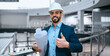 © Prostock-studio - A construction worker is smiling and giving a thumbs up at a building site. He holds blueprints and wears a hard hat. The construction area is busy with equipment and structures.