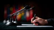 © Secrets inside - A close-up of a hand holding a pen poised to write on paper, with multiple microphones and blurred colorful flags in the background, suggesting a formal or official setting