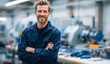 © Selvi - Smiling confident man with a well-groomed beard and short brown hair, wearing a blue work jacket, standing with arms crossed in an industrial manufacturing facility