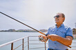 © TrueShot | Westend61 - Smiling man fishing on riverside deck wearing sunglasses and casual shirt