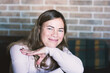 © dizfoto1973 - A smiling woman with light brown hair poses happily indoors, conveying warmth and approachability against a blurred brick background. Her genuine expression radiates positivity.