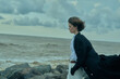 © SHOTPRIME STUDIO - Windy coastal scene of a woman on rocks by the sea, coat billowing behind, calm expression, looking toward the horizon under moody clouds, outdoor fashion mood with natural elements.