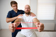 © Василь Івасюк - Physiotherapist guiding male patient with resistance band exercise for shoulder rehabilitation in clinic. Physical therapy session focused on injury recovery, strengthening, mobility improvement