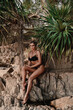 © blackday - Stylish woman wearing black lace lingerie poses on rocky tropical beach surrounded by palm trees