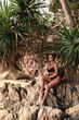 © blackday - Stylish woman wearing black lace lingerie poses on rocky tropical beach surrounded by palm trees